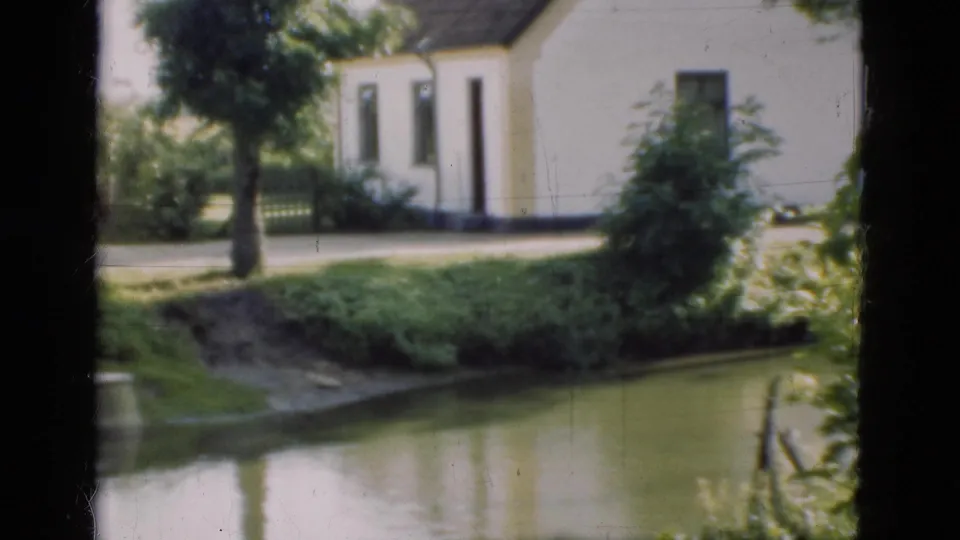 1975 Denmark 8mm frame showing a white waterside building, tree, green bushes, and water reflecting the bank.
