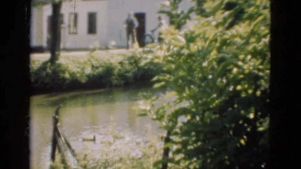 1975 Denmark 8mm view of a white house near water, green foliage, and distant people standing by the building.