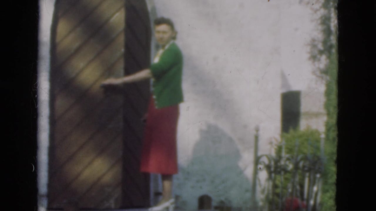Woman in a green cardigan and red skirt stands beside a large wooden door.