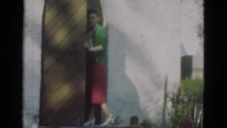 The woman is partly obscured by the door, with her red skirt and light shoes visible in the soft 8mm frame.
