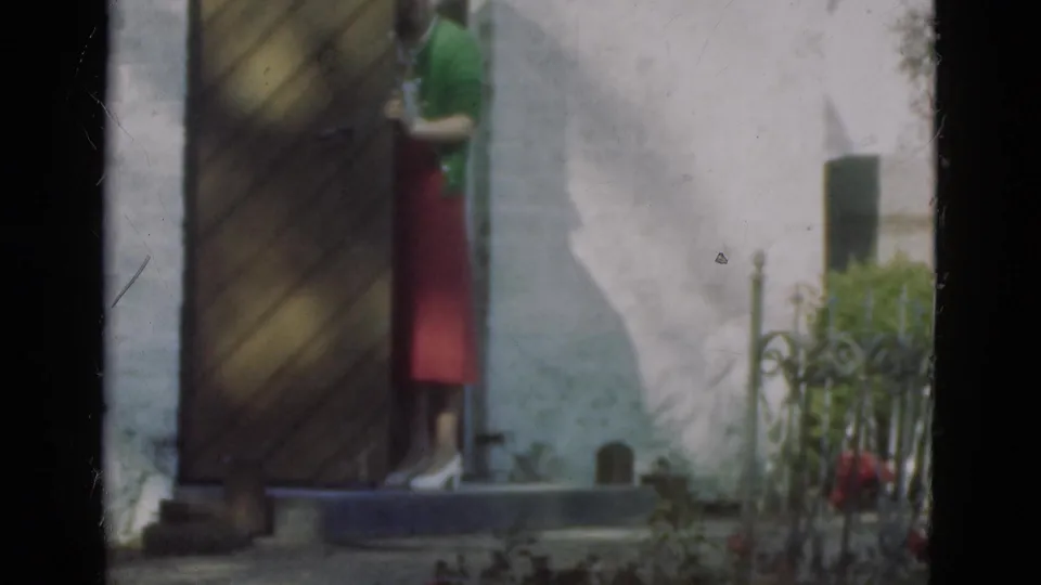 A woman in a green cardigan and red skirt stands mostly behind a large wooden door in Danish 1975 home-movie footage.