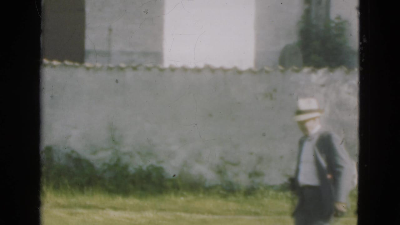 A man in a brimmed hat walks across grass near a pale stone wall.