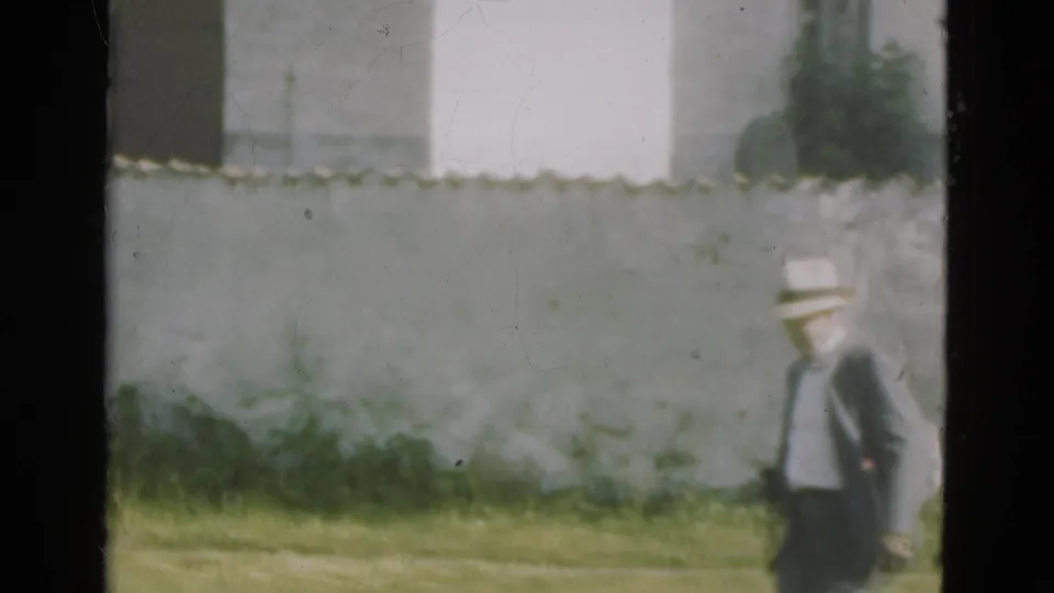 In Denmark 1975 home-movie footage, a suited man in a brimmed hat moves across grass beside a pale stone wall.