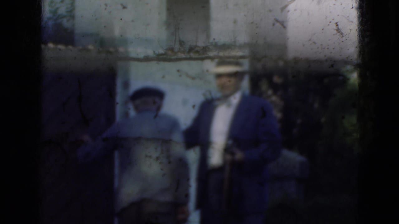 Two adults appear near a stone doorway in Denmark, with the hat-wearing man holding a small camera at his side.