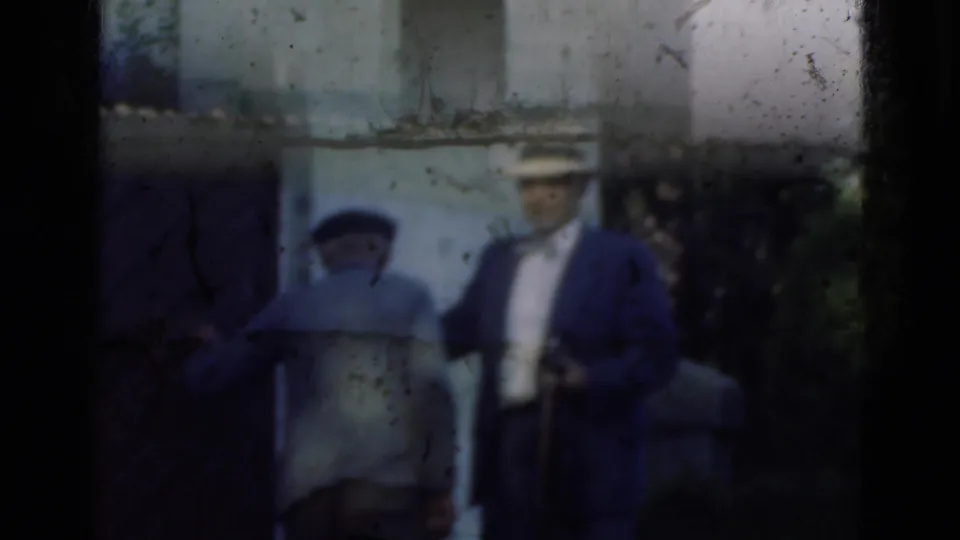 Two adults appear near a stone doorway in Denmark, with the hat-wearing man holding a small camera at his side.