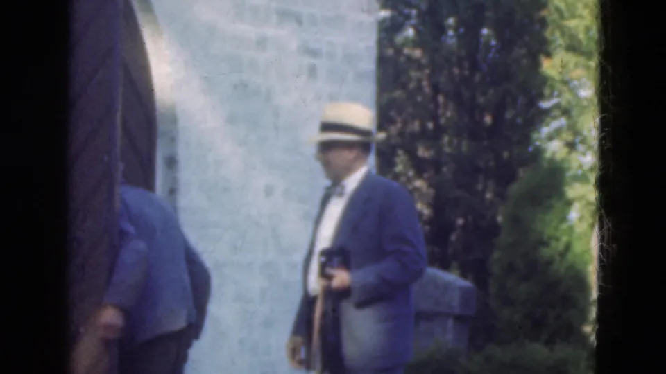The suited visitor in a brimmed hat holds a camera while standing beside a pale stone entrance and dark doorway edge.