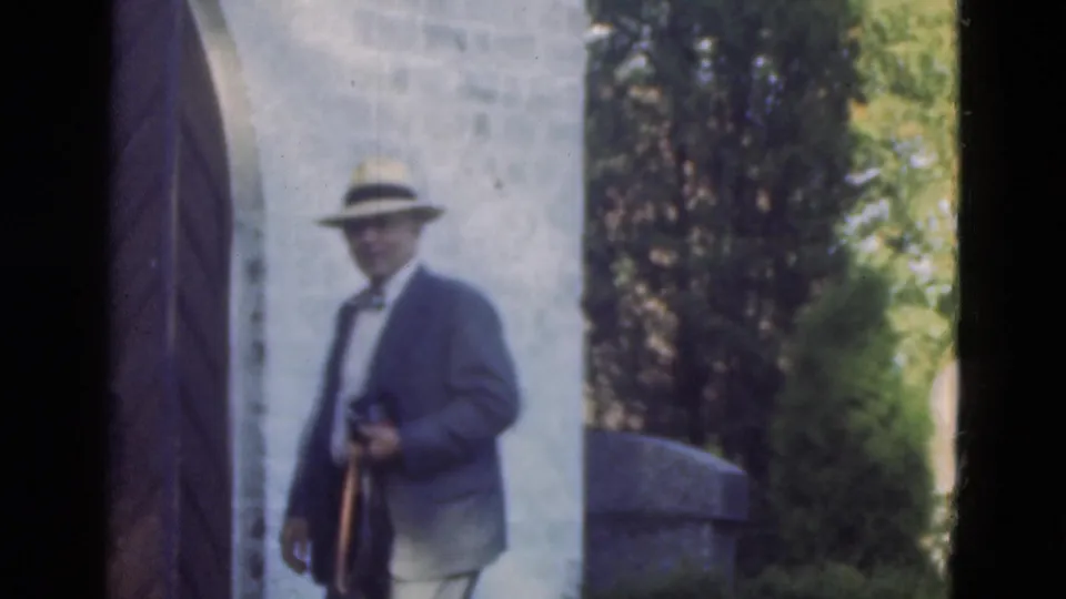 A suited man in a brimmed hat stands near a stone wall and greenery, holding a camera in Denmark 1975 footage.