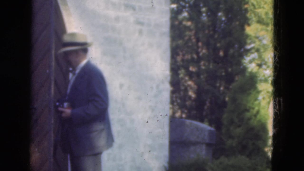 Man in a fedora stands near a garden wall with greenery behind him.