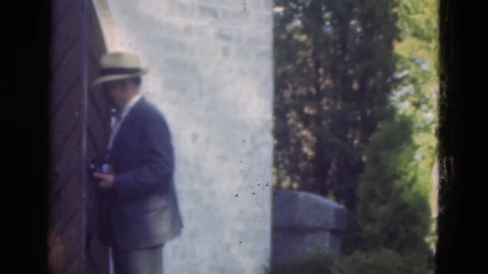 In Denmark 1975 home-movie footage, a man in a fedora and dark jacket appears beside a pale wall and garden plants.