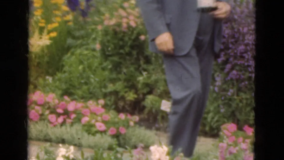 A suited man holding a small camera walks through a flower garden in the 1975 Denmark home-movie clip.