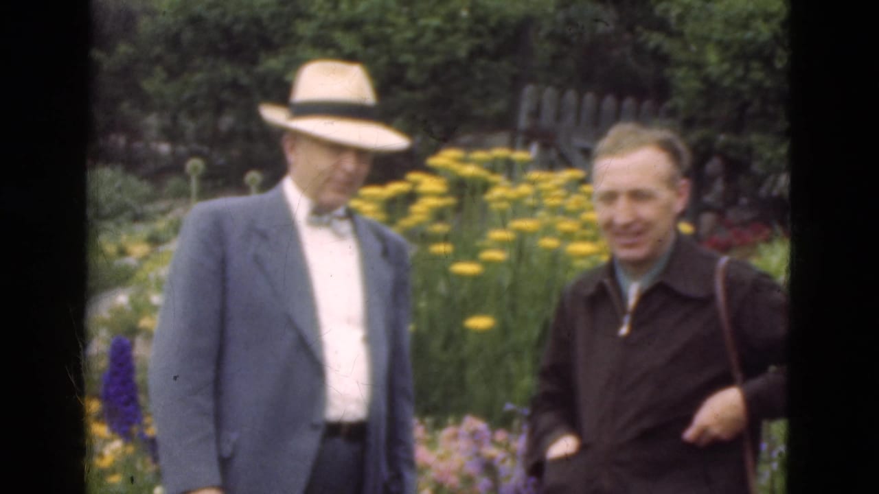 1975 Denmark 8mm frame of two adult men standing among yellow flowers, with one in a tan fedora, bow tie, and blue sport coat.