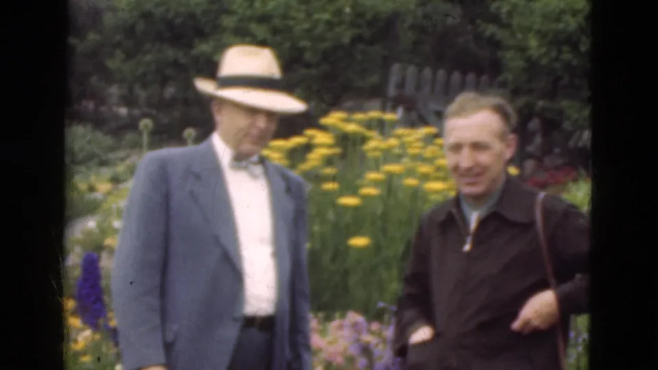1975 Denmark 8mm frame of two adult men standing among yellow flowers, with one in a tan fedora, bow tie, and blue sport coat.