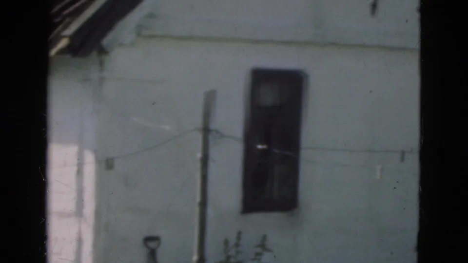 1975 Denmark 8mm frame showing a white rural building corner with a dark opening, tiled roof edge, and a line or wire across the garden area.