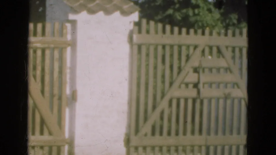 1975 Denmark 8mm frame of a pale wooden gate beside a white wall, seen through a dark window frame with greenery near the top edge.