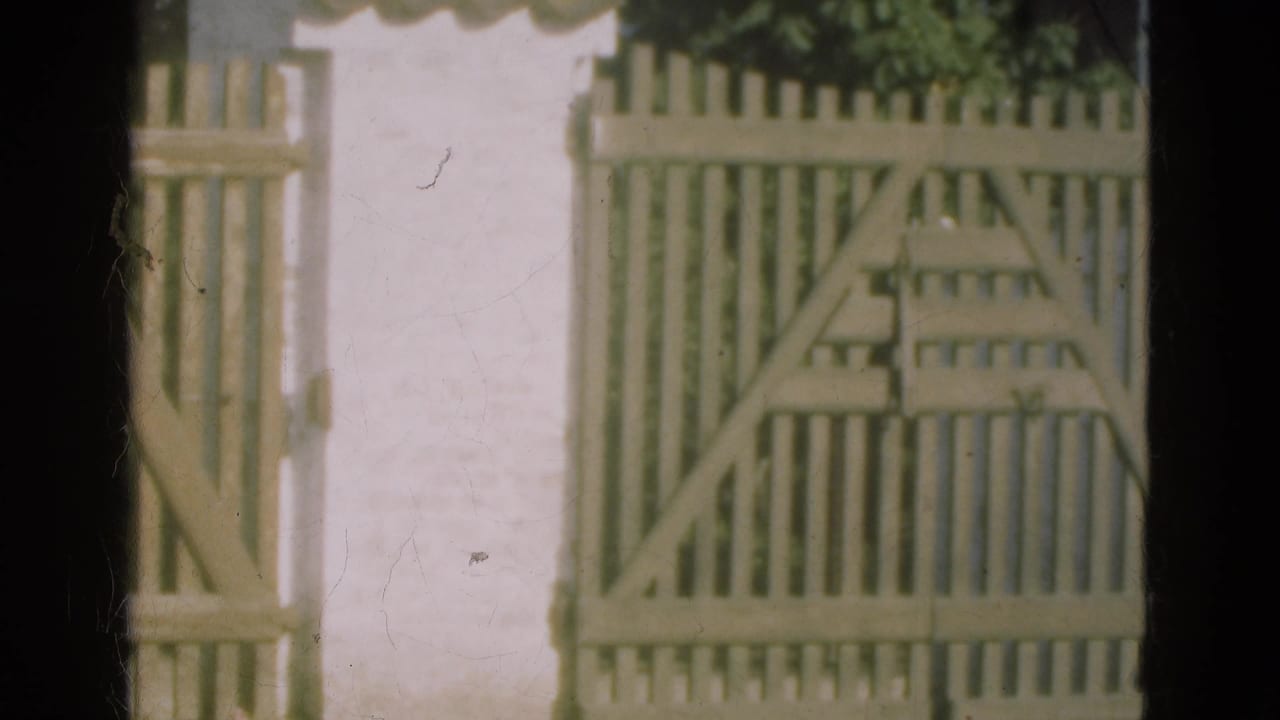 1975 Denmark 8mm frame showing a pale wooden gate and picket fence against a white stucco wall with soft focus and film wear.