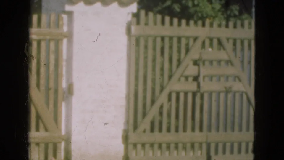1975 Denmark 8mm frame showing a pale wooden gate and picket fence against a white stucco wall with soft focus and film wear.