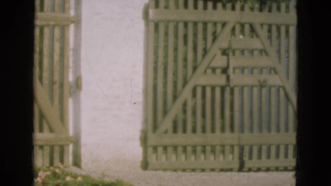1975 Denmark 8mm frame with a closed pale wooden gate, vertical fence slats, and white stucco wall in soft vintage color.