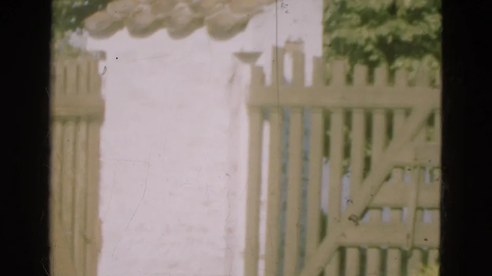 1975 Denmark 8mm frame showing the wooden gate partly swung open near a white stucco wall and leafy greenery.