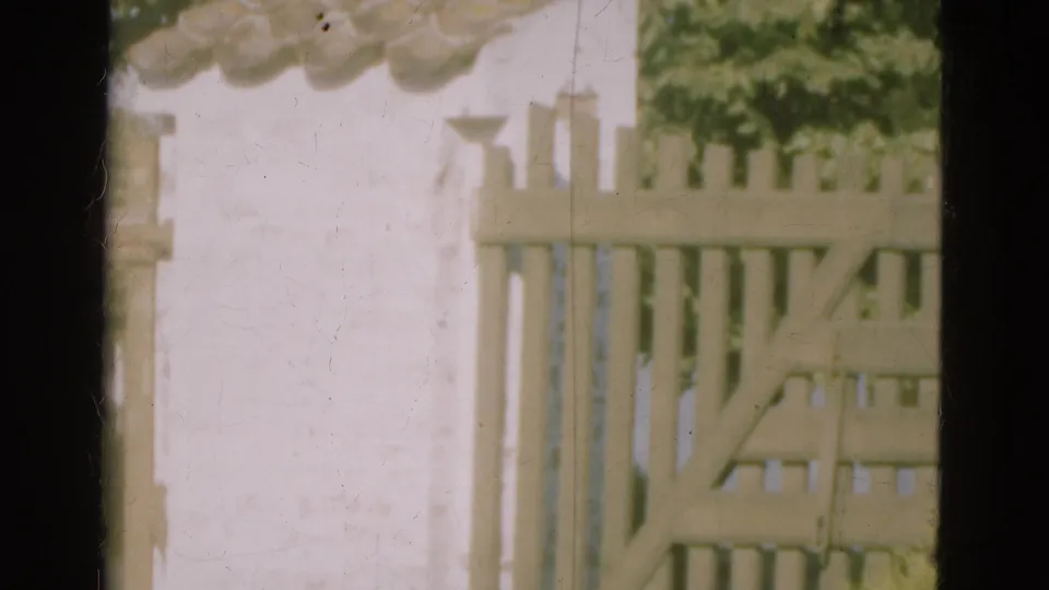 1975 Denmark 8mm frame with the gate open near a pale fence and white stucco wall, showing visible dust and soft focus.