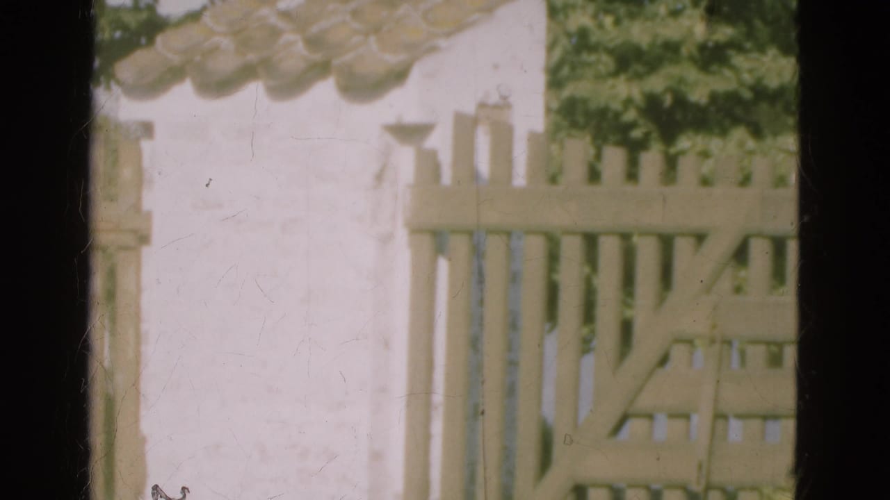1975 Denmark 8mm frame showing a sunlit white wall, clay roof tiles, wooden picket fence, and greenery beyond.