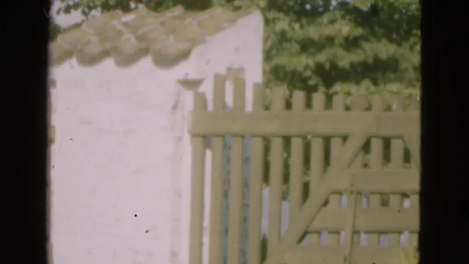 1975 Denmark home-movie frame with a white stucco surface, wooden gate or fence slats, roof tiles, and background foliage.