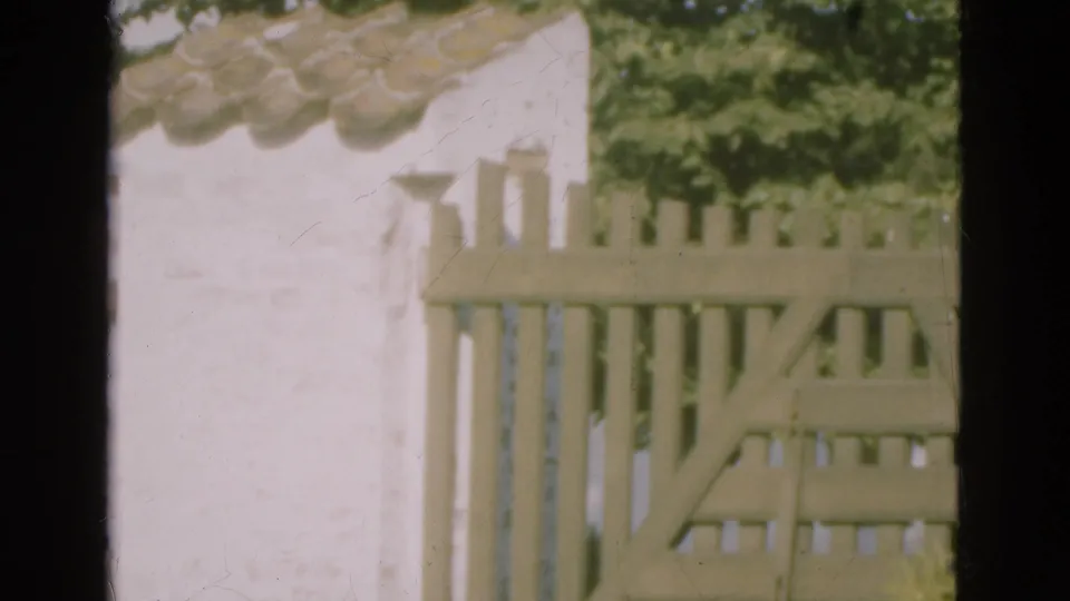 Faded 8mm frame from Denmark showing a sunlit wooden fence, white wall, roof tiles, and leafy greenery in the background.