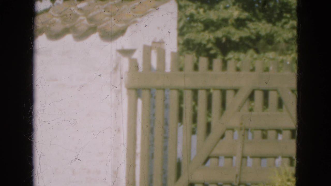 1975 Denmark 8mm frame showing a pale slatted wooden gate, white wall, tiled roofline, and green foliage in soft focus.