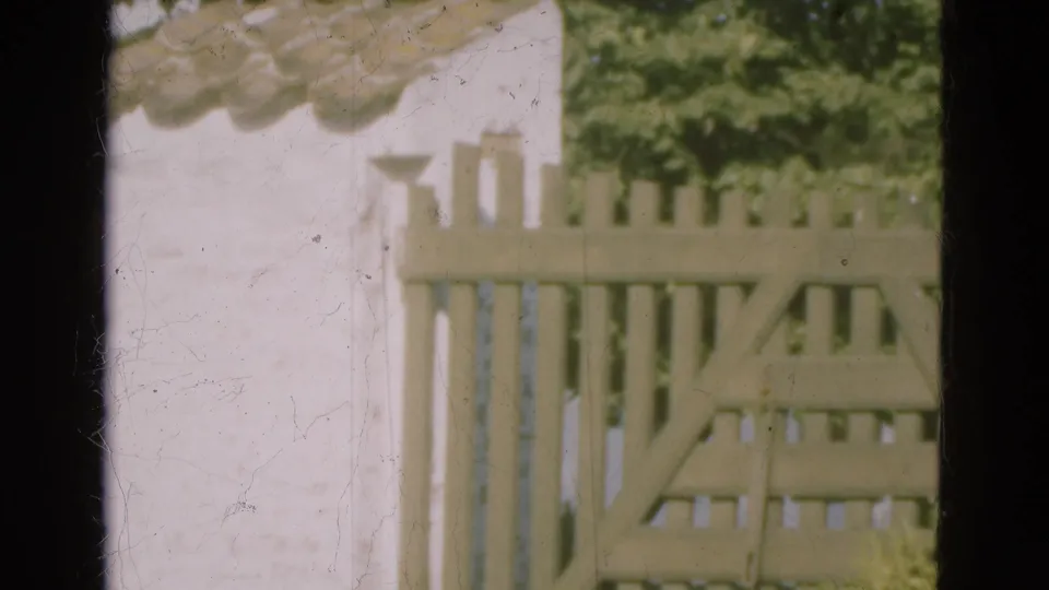 1975 Denmark 8mm frame showing a pale slatted wooden gate, white wall, tiled roofline, and green foliage in soft focus.