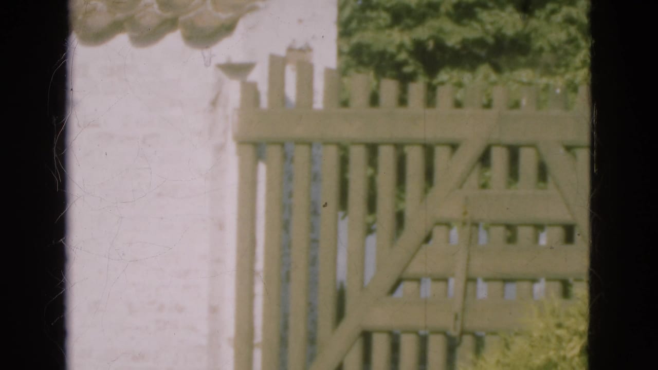 1975 Denmark 8mm frame of the slatted gate and white wall, with greenery behind and dark film-frame edges visible.