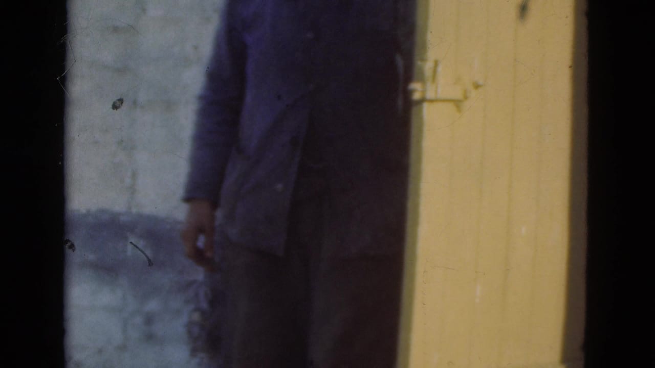 Man in a blue jacket standing beside a pale wall and yellow-brown wooden door.