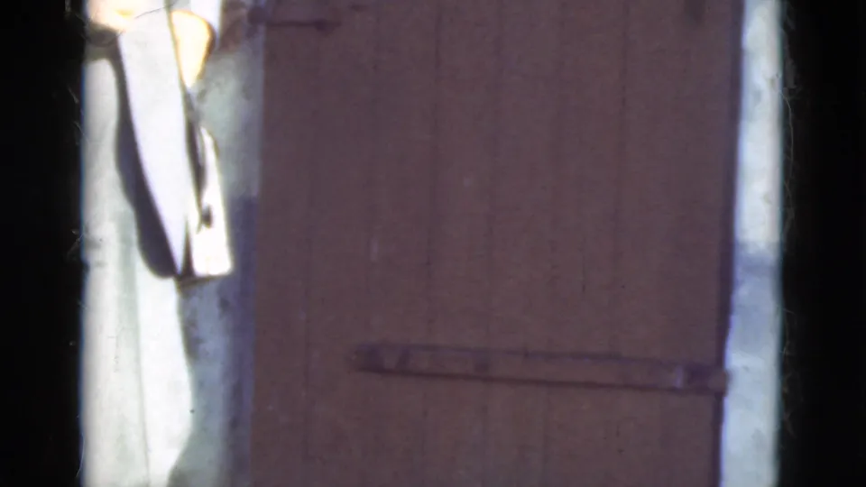 A cropped person stands beside the closed or nearly closed brown wooden door, with the metal latch visible in soft 8mm color.