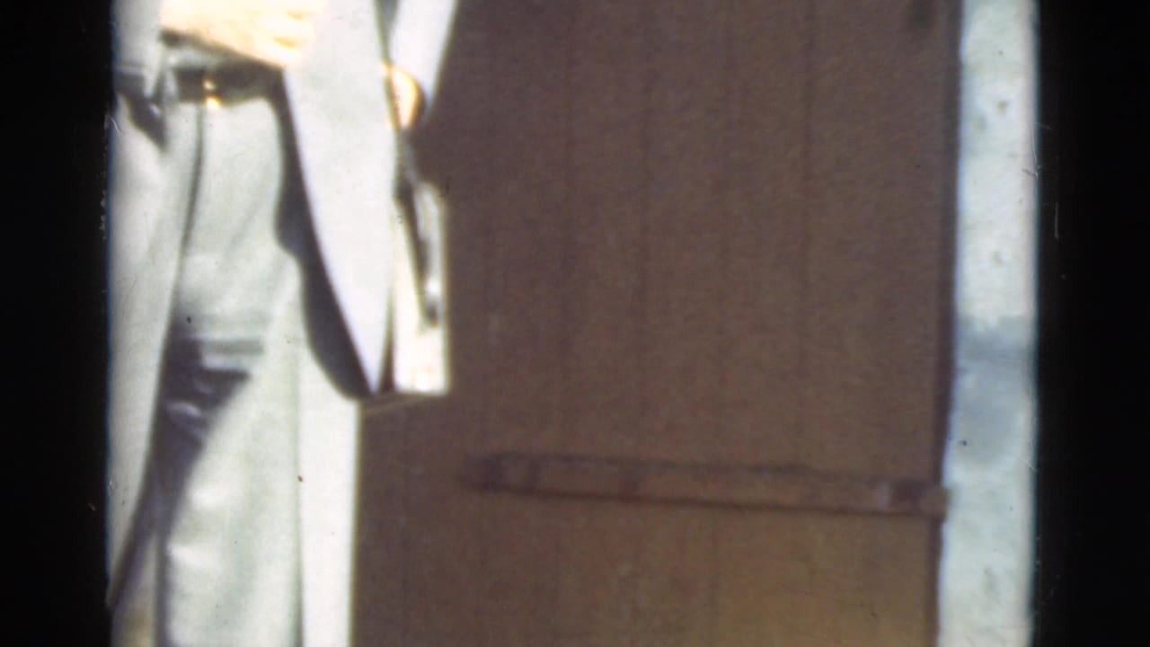 Partial person in light clothing beside a wooden door, seen through dark window framing.
