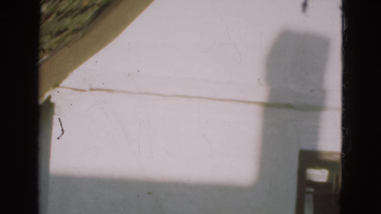 1975 Danish home-movie frame of a pale house wall, sloped roof edge, and a large shadow crossing the exterior.