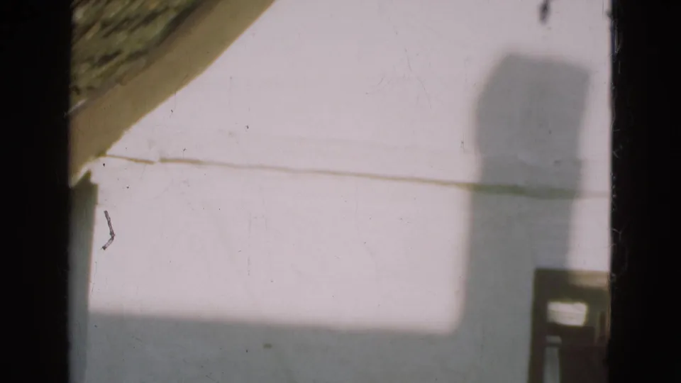 1975 Danish home-movie frame of a pale house wall, sloped roof edge, and a large shadow crossing the exterior.
