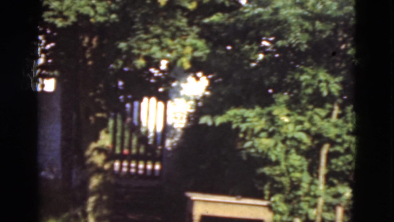 1975 Denmark 8mm frame showing a shaded path, foliage, and a wooden gate near a white exterior wall.