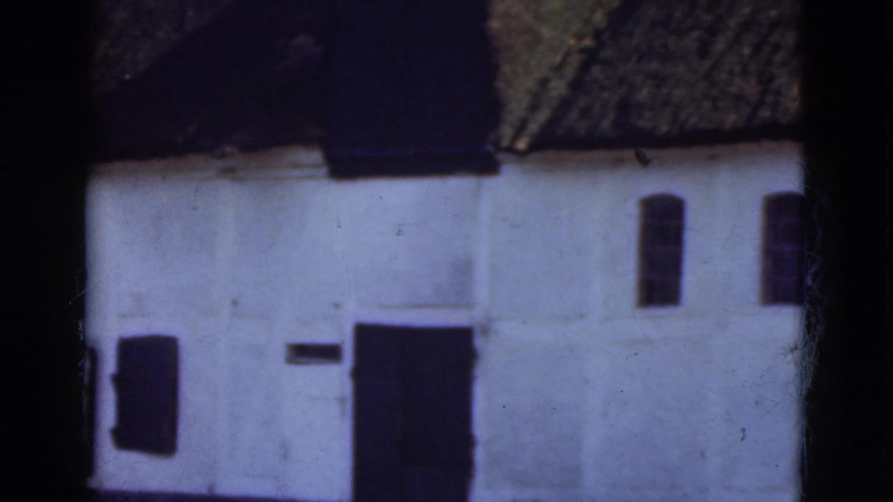 1975 Denmark 8mm frame of a whitewashed thatched-roof building with dark openings and trees nearby.