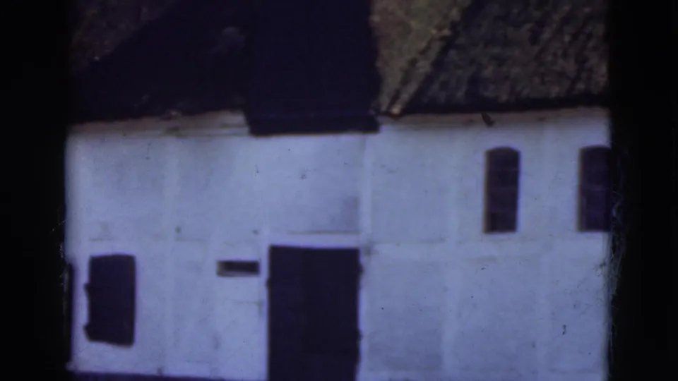 1975 Denmark 8mm frame of a whitewashed thatched-roof building with dark openings and trees nearby.
