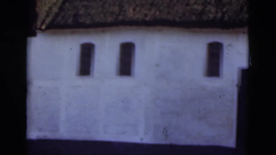 1975 Denmark 8mm frame showing the side of a white thatched-roof house with several dark narrow windows.