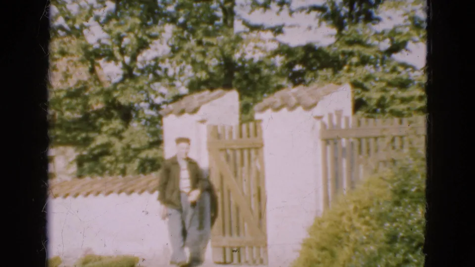 1975 Denmark 8mm frame with a man at a wooden gate before a white thatched-roof house and leafy trees.