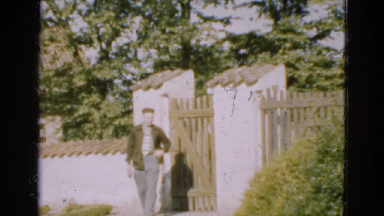 Man stands near a white garden wall and wooden gate under leafy trees.