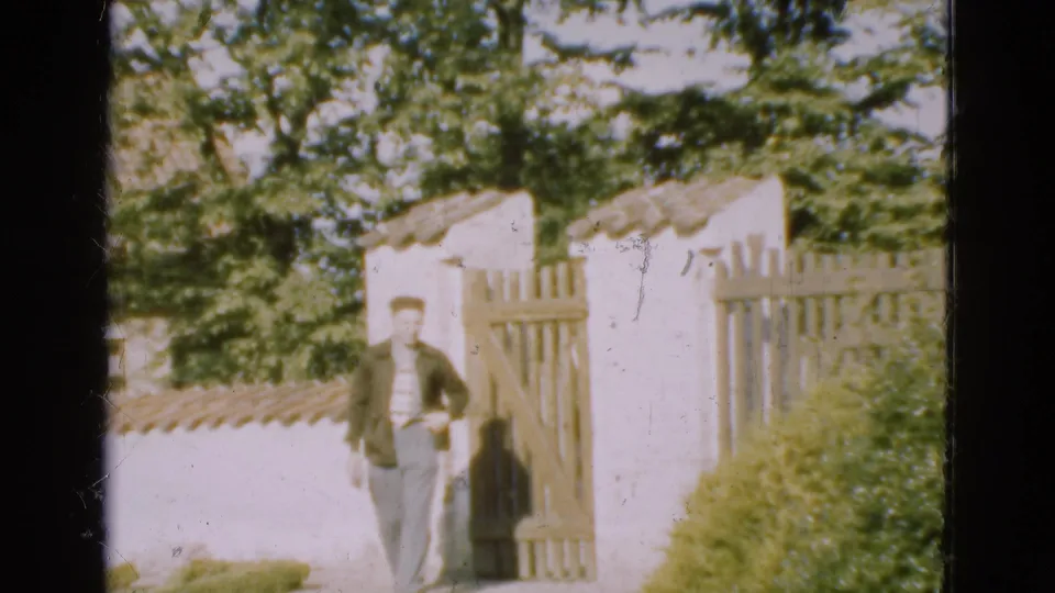 In 1975 Denmark 8mm footage, a man in a dark jacket stands near a white garden entrance with a wooden gate and trees overhead.