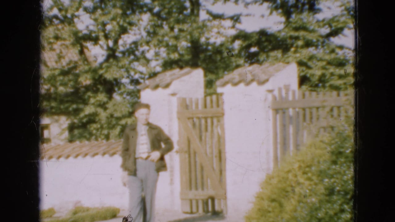 The man walks toward the camera from a wooden gate set into a white-walled garden entrance in Denmark, 1975.