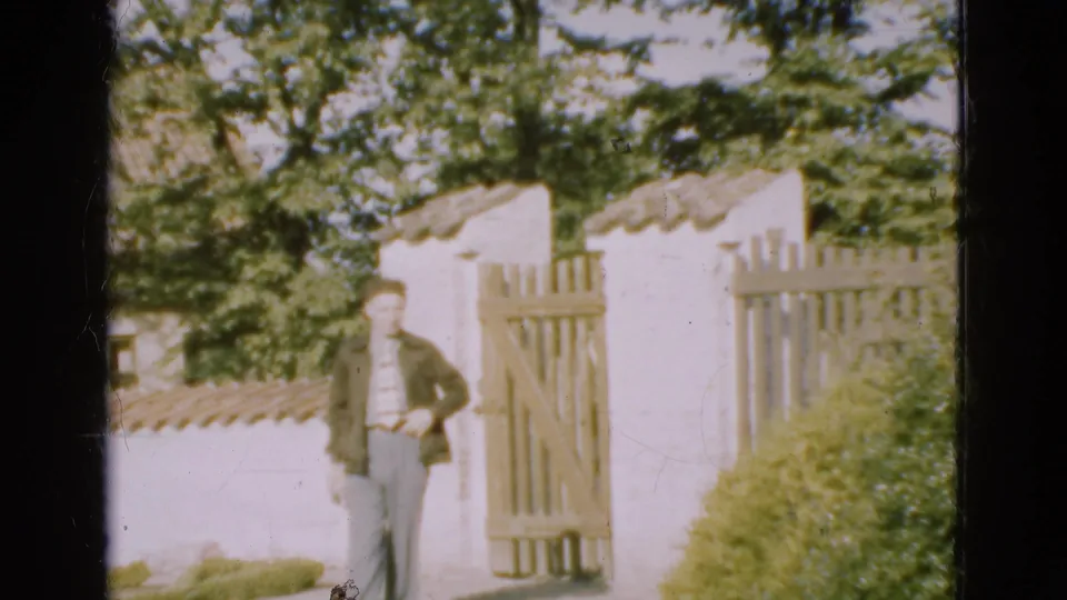 The man walks toward the camera from a wooden gate set into a white-walled garden entrance in Denmark, 1975.