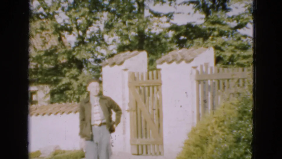 A closer 8mm frame shows the man nearing the camera, with the wooden gate, white wall, trees, and garden shrubs behind him.