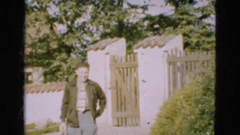 At the end of the clip, the man is closest to camera, carrying a small object with the white wall and wooden gate still visible behind him.