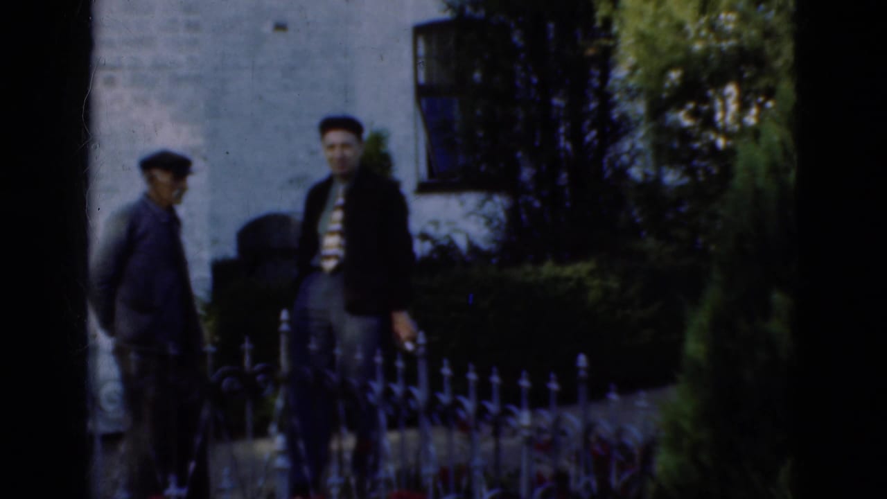 In Denmark 1975 home-movie footage, two adult men stand by an iron fence in front of a white brick building with shrubs nearby.