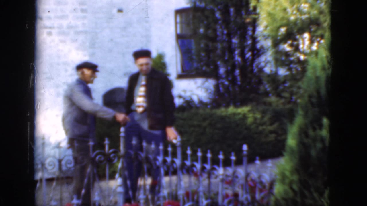 In the supported Denmark 1975 setting, two adult men appear to shake hands beside an ornate iron fence and white brick exterior.