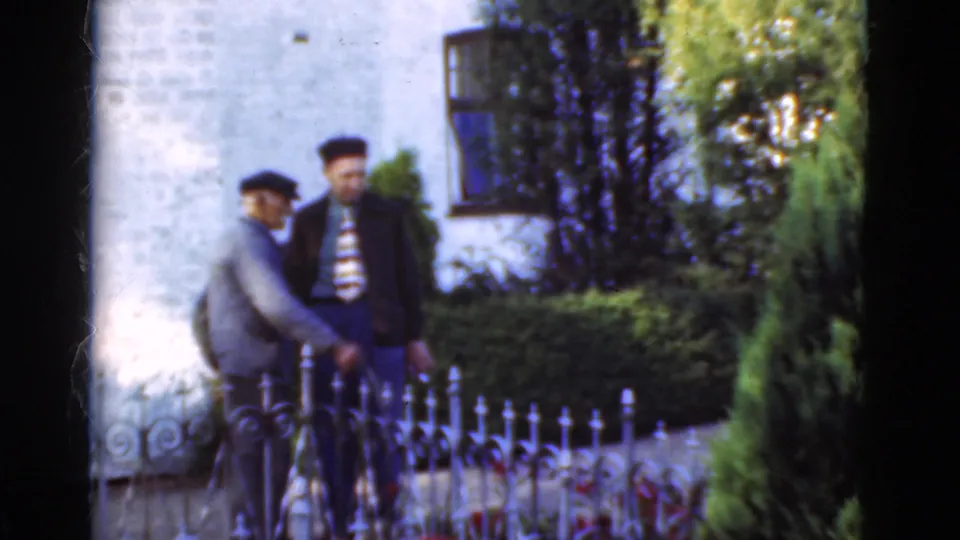 Two adult men in a 1975 Denmark home-movie scene stand close by an iron fence, with greenery and a white brick wall behind them.