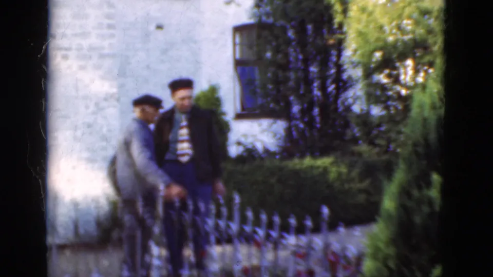 The Denmark 1975 footage shows two men near an ornate iron fence, with one looking down and the other standing against the white brick exterior.