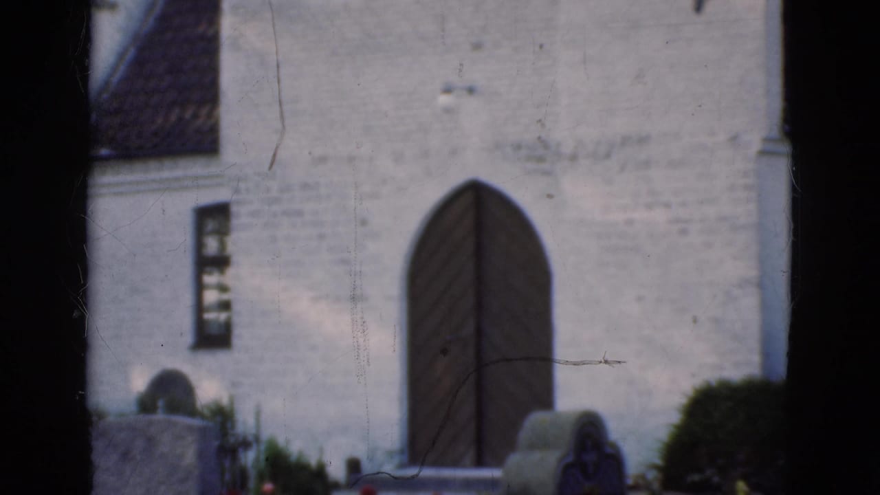 White church wall with arched wooden door, gravestones, hedge, and dark tree edges.